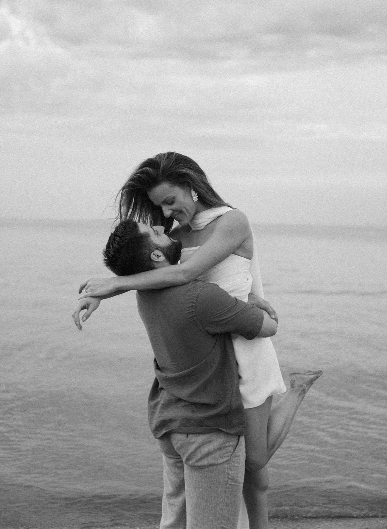 Black and white photo of a couple embracing on a beach with a cloudy sky.